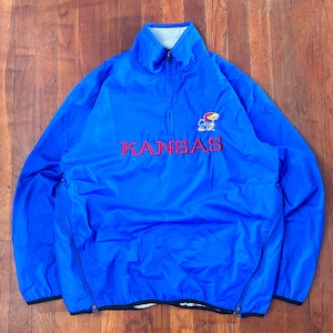 May include: A blue pullover jacket with a high collar and a half-zip closure. The word "KANSAS" is embroidered in red across the chest, with a Jayhawk logo above. The jacket has side pockets with zippers and a white lining.