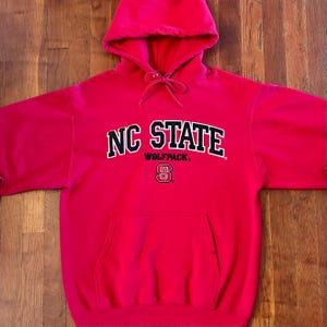 Puede incluir: Una sudadera con capucha de color rojo brillante que presenta el texto NC STATE en letras negras con bordes blancos, la palabra WOLFPACK en letras negras m&aacute;s peque&ntilde;as y un logotipo de NC State en rojo y blanco. Incluye bolsillo frontal y cordones.