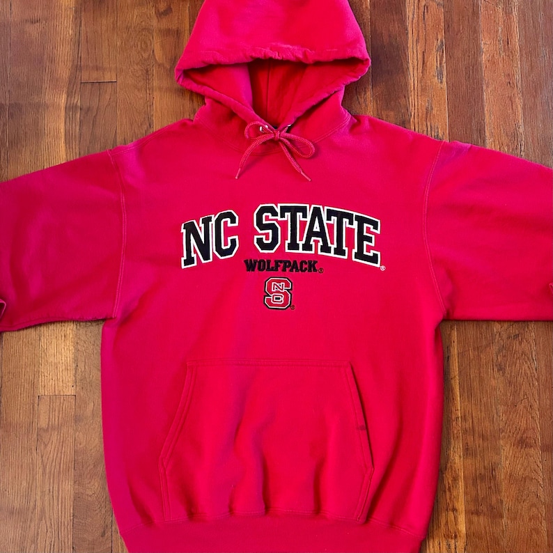 Puede incluir: Una sudadera con capucha de color rojo brillante que presenta el texto NC STATE en letras negras con bordes blancos, la palabra WOLFPACK en letras negras m&aacute;s peque&ntilde;as y un logotipo de NC State en rojo y blanco. Incluye bolsillo frontal y cordones.