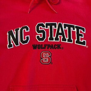 Puede incluir: Una sudadera con capucha de color rojo brillante que presenta el texto NC STATE en letras negras grandes con bordes blancos. Debajo, la palabra WOLFPACK est&aacute; impresa en negro, seguida de un emblema con el logo NC en rojo y blanco.