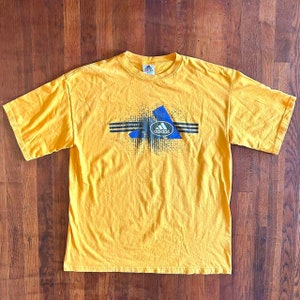May include: A yellow t-shirt with a blue and black Adidas logo and graphic design. The logo is surrounded by a black circle with the word "adidas" in white.