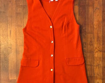 May include: A sleeveless, orange vest with a V-neck and four gold-colored buttons down the front. The vest has two front pockets and is made of a textured fabric. The garment is displayed on a wooden surface.