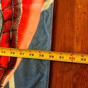 May include: A vibrant beach towel with a superhero theme, showcasing red, blue, and orange colours. The towel measures approximately 81 cm in length, as indicated by a yellow tape measure. The towel is on a wooden surface.