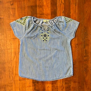 May include: A light blue denim top with short sleeves and a round neckline. The top features yellow and black embroidery around the neckline and on the shoulders.