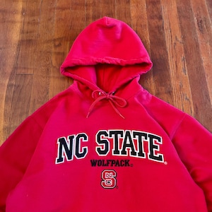 Puede incluir: Una sudadera con capucha de color rojo brillante que presenta el texto NC STATE en letras grandes de color negro con bordes blancos. Debajo del texto, la palabra WOLFPACK est&aacute; impresa en negro, seguida de un peque&ntilde;o logo NC en rojo, blanco y negro.