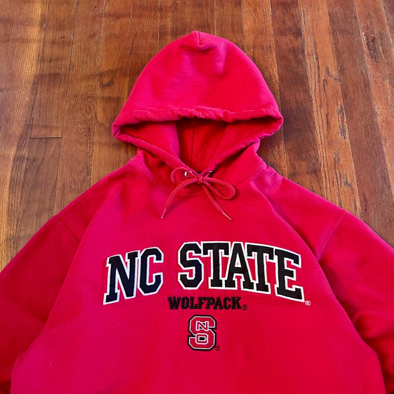 Puede incluir: Una sudadera con capucha de color rojo brillante que presenta el texto NC STATE en letras grandes de color negro con bordes blancos. Debajo del texto, la palabra WOLFPACK est&aacute; impresa en negro, seguida de un peque&ntilde;o logo NC en rojo, blanco y negro.
