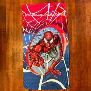 May include: A rectangular beach towel featuring Spider-Man in a dynamic pose, set against a web design and a blue and red background. The words "SPIDER-MAN 2" are printed at the top.