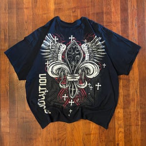 May include: Black t-shirt featuring a fleur-de-lis design with white wings, crosses, and the word "evolution" in white. The graphic has red accents. The shirt is laid flat on a wooden surface.