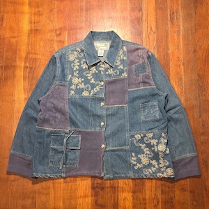 May include: A long-sleeve denim jacket featuring a patchwork design with blue denim panels, solid purple fabric sections, and cream-colored floral embroidery. The jacket includes a button-down front, a collar, and a square pocket with a drawstring.