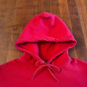 Puede incluir: Una sudadera con capucha de color rojo brillante s&oacute;lido que presenta una construcci&oacute;n de capucha de doble capa y cordones tejidos rojos a juego atados en un lazo en el centro del cuello. La prenda est&aacute; hecha de tela lisa con costuras visibles.