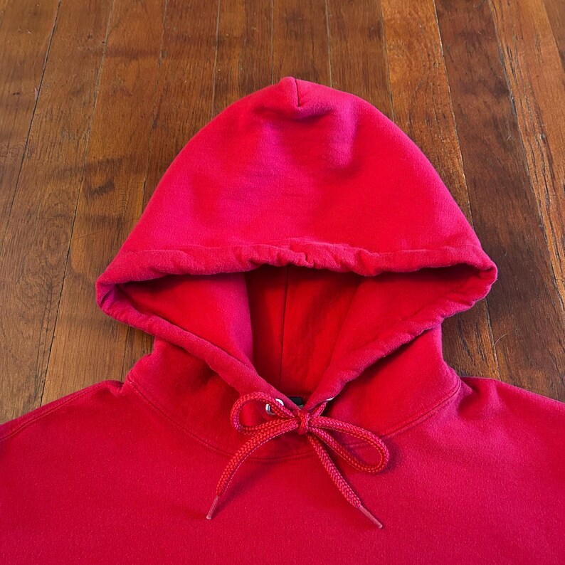 Puede incluir: Una sudadera con capucha de color rojo brillante s&oacute;lido que presenta una construcci&oacute;n de capucha de doble capa y cordones tejidos rojos a juego atados en un lazo en el centro del cuello. La prenda est&aacute; hecha de tela lisa con costuras visibles.