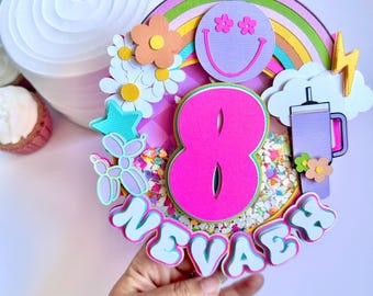 May include: A pink and white cake topper with a rainbow, a smiley face, a number 8, and the name "Nevaeh".