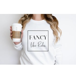 May include: White sweatshirt with black text that reads "FANCY like Reba" in a square frame.
