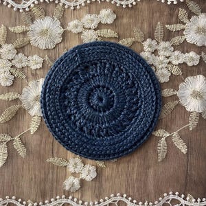 Puede incluir: Un posavasos circular de ganchillo azul marino con un dise&ntilde;o en espiral. El posavasos est&aacute; hecho de un hilo grueso y tiene un aspecto texturizado. El posavasos tiene aproximadamente 10 cm de di&aacute;metro.