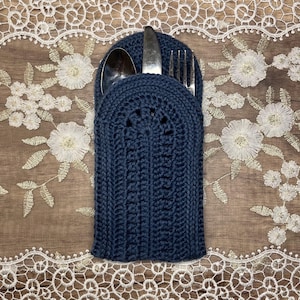 May include: A navy blue, crocheted cutlery holder with a spoon, knife, and fork inside. The holder has a textured, ribbed design and a decorative circular pattern near the top. The background features a wooden surface and a white lace doily.
