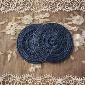 Puede incluir: Dos posavasos de ganchillo azul marino con un dise&ntilde;o circular. Los posavasos est&aacute;n hechos de un hilo grueso y tienen un aspecto texturizado. Se colocan sobre una superficie marr&oacute;n claro con encaje floral blanco y dorado.