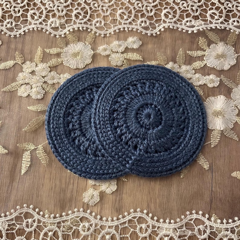 Puede incluir: Dos posavasos de ganchillo azul marino con un dise&ntilde;o circular. Los posavasos est&aacute;n hechos de un hilo grueso y tienen un aspecto texturizado. Se colocan sobre una superficie marr&oacute;n claro con encaje floral blanco y dorado.