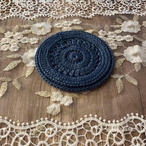Puede incluir: Un conjunto de tres posavasos de ganchillo azul marino. Los posavasos son circulares con un dise&ntilde;o en espiral en el centro. Los posavasos est&aacute;n sobre una superficie con bordados florales blancos y dorados.