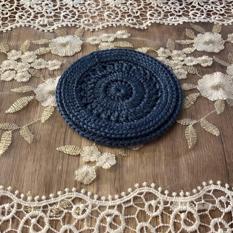 Puede incluir: Un conjunto de tres posavasos de ganchillo azul marino. Los posavasos son circulares con un dise&ntilde;o en espiral en el centro. Los posavasos est&aacute;n sobre una superficie con bordados florales blancos y dorados.