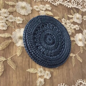 Puede incluir: Un posavasos redondo de ganchillo azul marino con un dise&ntilde;o en espiral. El posavasos est&aacute; hecho de hilo grueso y tiene una superficie texturizada. Est&aacute; sobre una superficie beige con bordados florales blancos.