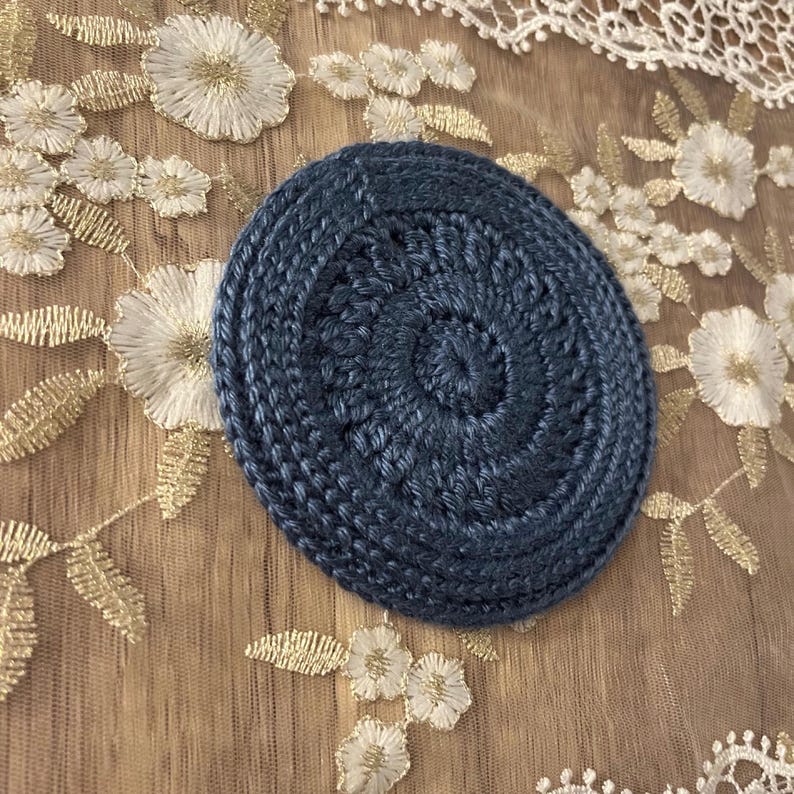 Puede incluir: Un posavasos redondo de ganchillo azul marino con un dise&ntilde;o en espiral. El posavasos est&aacute; hecho de hilo grueso y tiene una superficie texturizada. Est&aacute; sobre una superficie beige con bordados florales blancos.