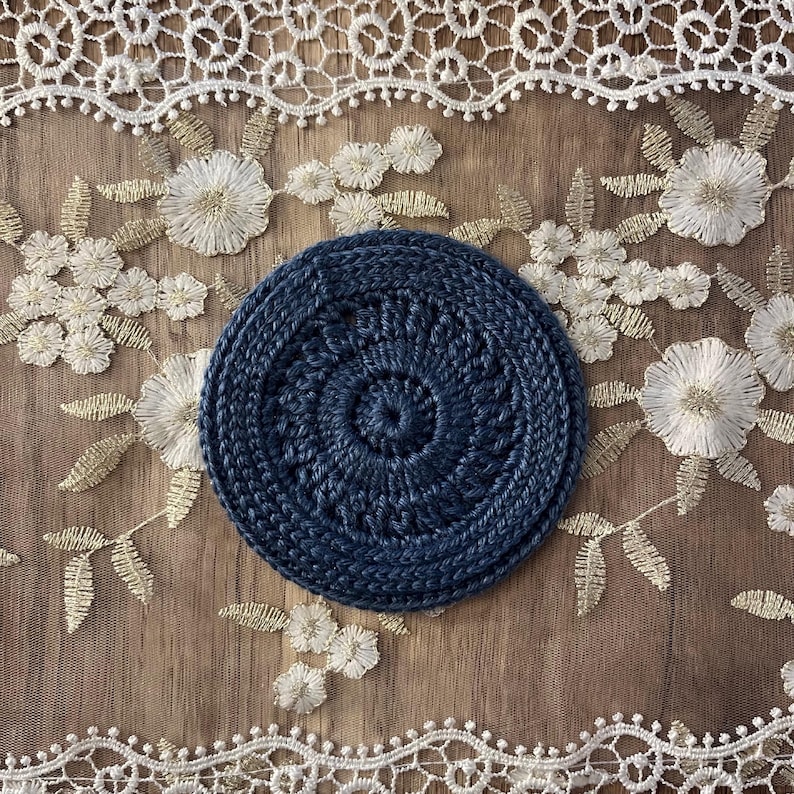 Puede incluir: Un posavasos circular de ganchillo azul marino con un dise&ntilde;o en espiral. El posavasos est&aacute; hecho de un hilo grueso y tiene un aspecto texturizado. El posavasos tiene aproximadamente 10 cm de di&aacute;metro.