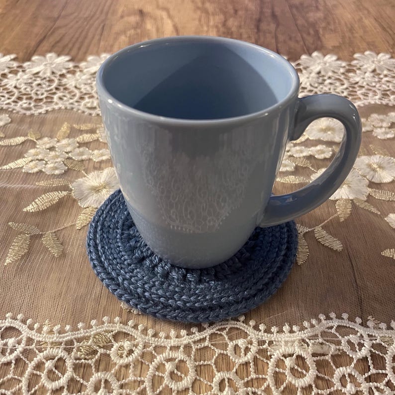 Puede incluir: Una taza de cer&aacute;mica azul claro con un asa curva descansa sobre un posavasos redondo de ganchillo azul marino. La taza y el posavasos est&aacute;n sobre una superficie de madera con un camino de mesa de encaje blanco.