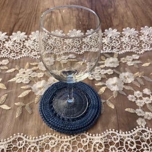 Puede incluir: Una copa de vino transparente descansa sobre un posavasos circular de punto azul marino. El posavasos est&aacute; hecho de hilo tejido apretado. El fondo presenta una superficie de madera con un camino de mesa de encaje floral blanco y dorado.