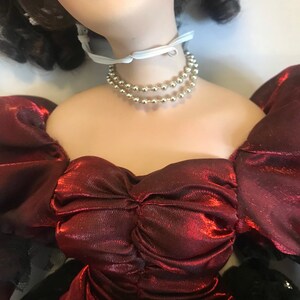 May include: A porcelain doll wearing a red satin dress with a ruffled neckline and a silver beaded necklace. The doll has long brown hair and is wearing a white ribbon around her neck.