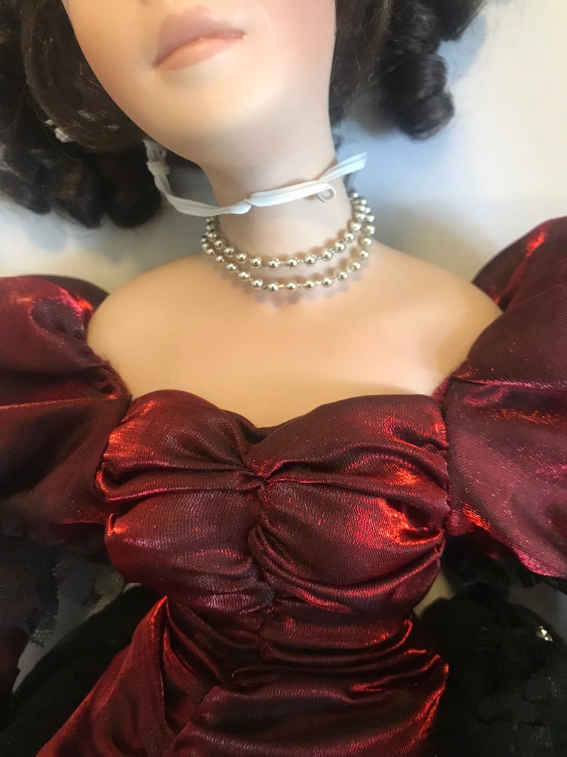 May include: A porcelain doll wearing a red satin dress with a ruffled neckline and a silver beaded necklace. The doll has long brown hair and is wearing a white ribbon around her neck.