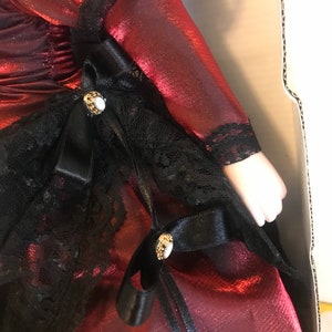 May include: A red satin dress with black lace trim and black satin ribbon bows. The dress has gold-colored buttons with white stones.