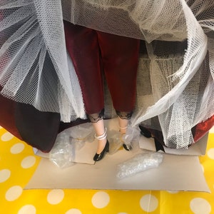 May include: A porcelain doll wearing a red and white dress with a white tulle skirt. The doll is wearing black shoes with white straps.