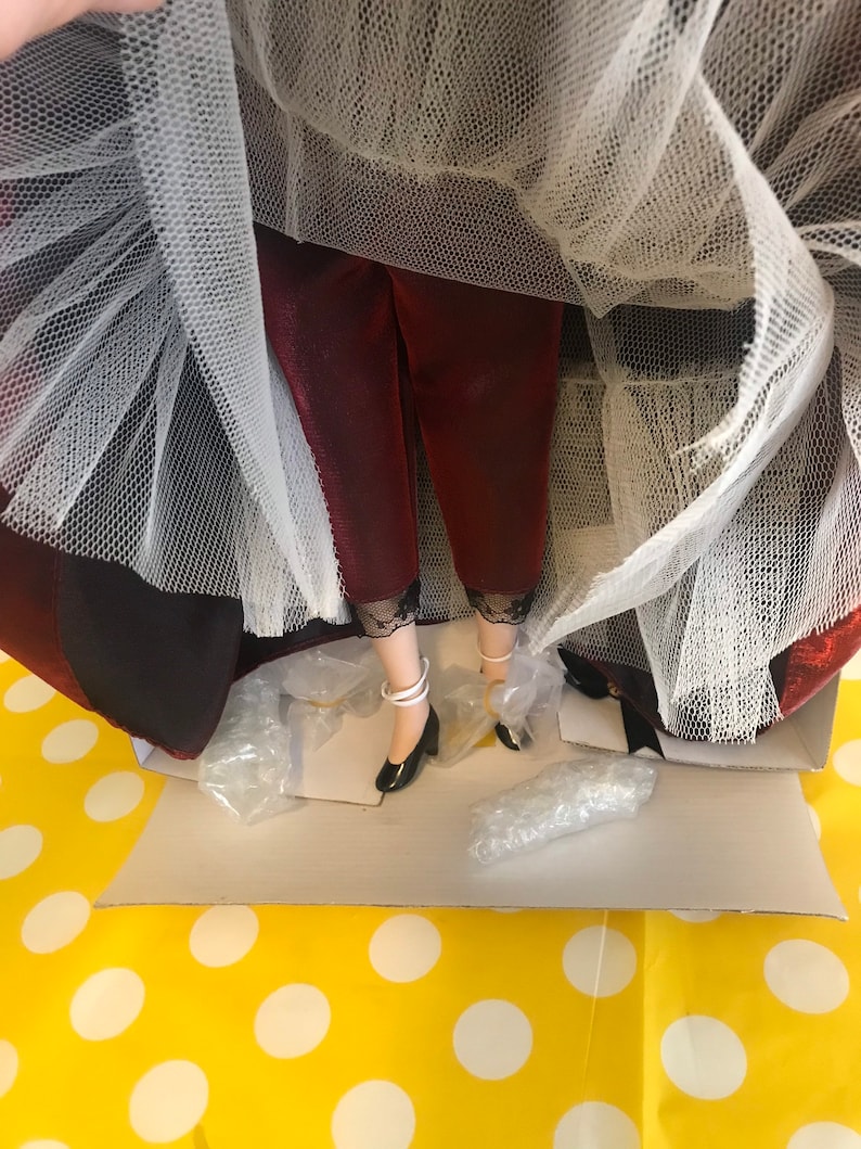 May include: A porcelain doll wearing a red and white dress with a white tulle skirt. The doll is wearing black shoes with white straps.