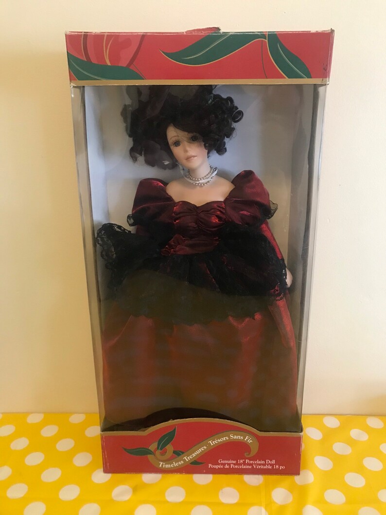 May include: A porcelain doll in a red and black dress with a black hat. The doll is in a box with the text "Timeless Treasures Tr&eacute;sors Sans Fir" and "Genuine 18" Porcelain Doll Poup&eacute;e de Porcelaine V&eacute;ritable 18 po".