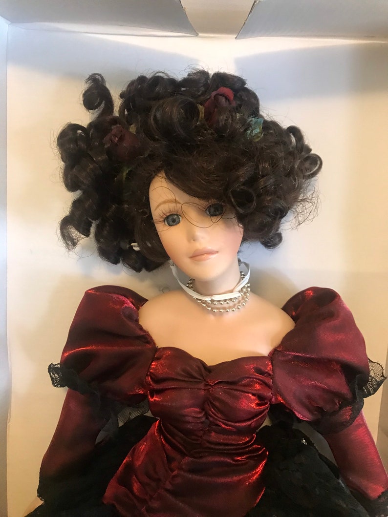 May include: A porcelain doll with dark brown curly hair, wearing a burgundy dress with a black lace trim and a pearl necklace.