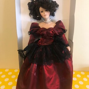May include: A porcelain doll wearing a burgundy satin dress with a black lace overlay and a pearl necklace. The doll has dark brown curly hair and brown eyes.