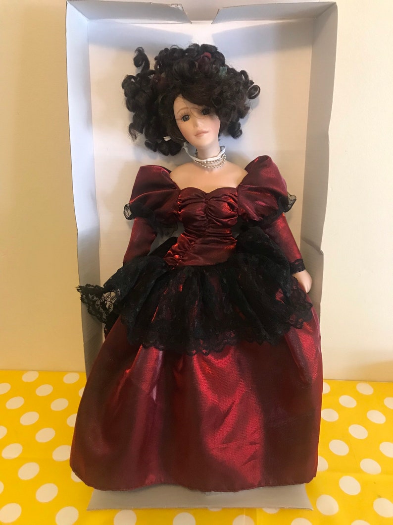 May include: A porcelain doll wearing a burgundy satin dress with a black lace overlay and a pearl necklace. The doll has dark brown curly hair and brown eyes.