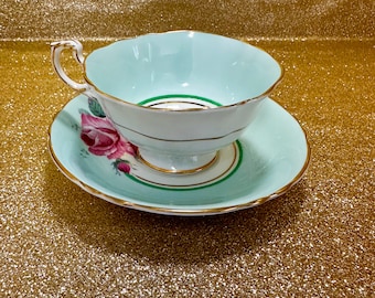 May include: A vintage teacup and saucer set. The teacup and saucer are light blue with gold trim. The saucer features a pink rose design. The teacup has a scalloped edge. The set is on a gold glitter background.