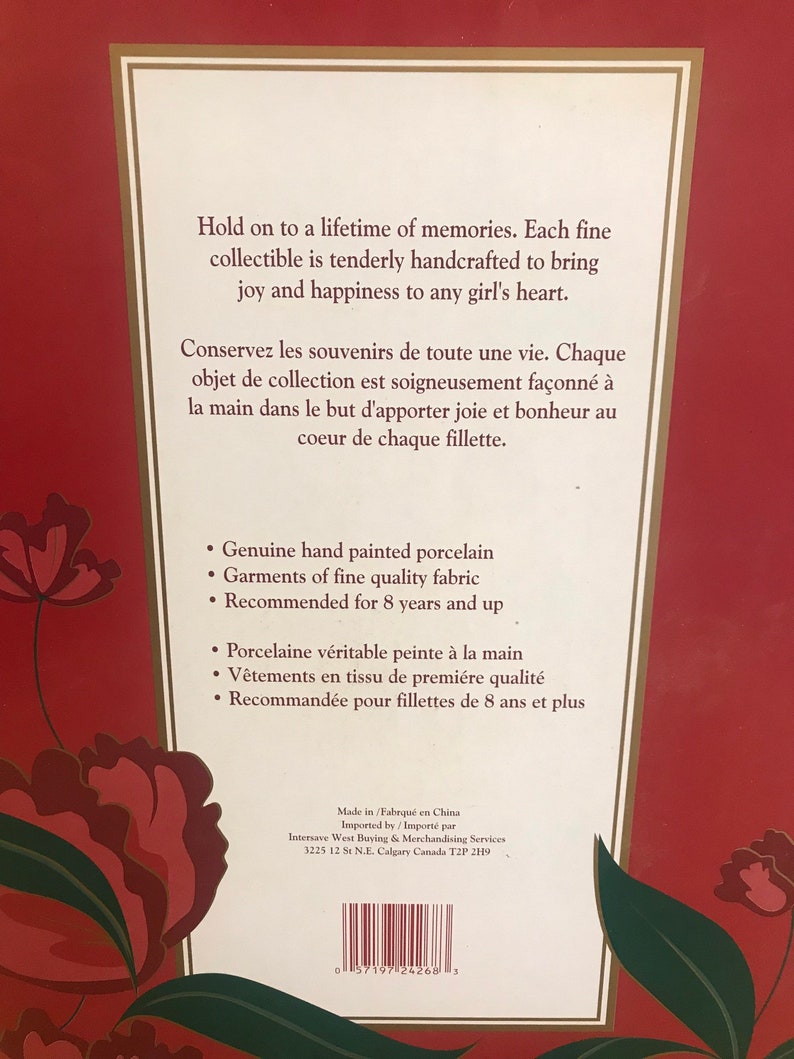 May include: Back of a box for a collectible doll. The box is red with a floral design. The text on the box reads "Hold on to a lifetime of memories. Each fine collectible is tenderly handcrafted to bring joy and happiness to any girl's heart." The box also includes a list of features, including "Genuine hand painted porcelain", "Garments of fine quality fabric", and "Recommended for 8 years and up".