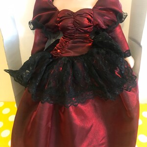 May include: A porcelain doll wearing a burgundy satin dress with a black lace overlay and a pearl necklace.