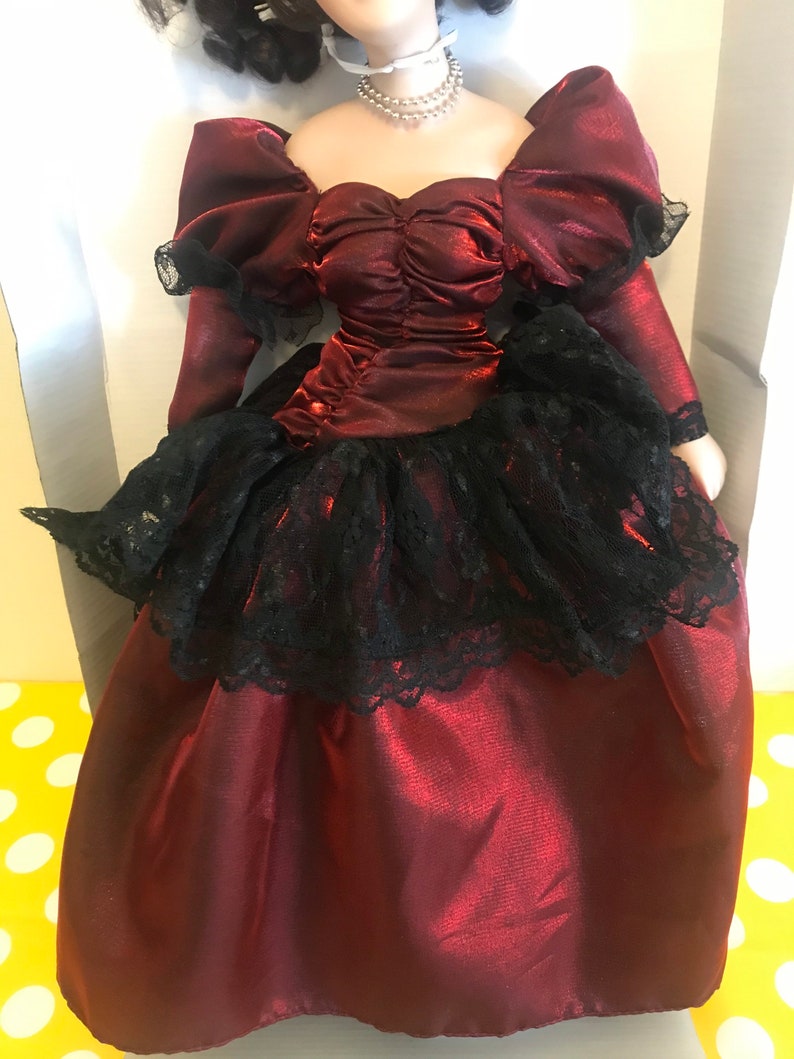May include: A porcelain doll wearing a burgundy satin dress with a black lace overlay and a pearl necklace.