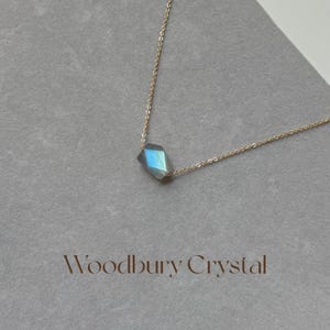 May include: A delicate gold chain necklace with a single, faceted, iridescent blue-grey gemstone pendant. The pendant is shaped like a diamond and hangs from the center of the chain.