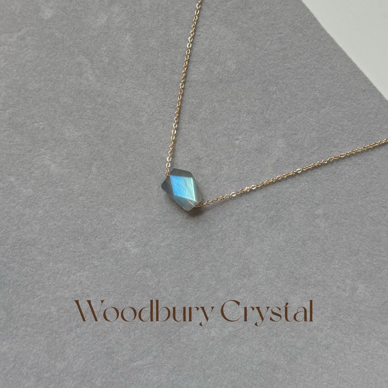 May include: A delicate gold chain necklace with a single, faceted, iridescent blue-grey gemstone pendant. The pendant is shaped like a diamond and hangs from the center of the chain.