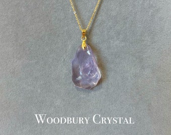 May include: A delicate gold chain necklace with a light purple crystal pendant. The crystal is irregularly shaped and has a translucent appearance. The necklace is labeled "Woodbury Crystal".