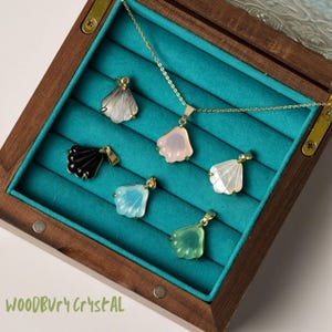 May include: A wooden box with a teal velvet interior displays a collection of six shell-shaped pendants. The pendants are made of various materials, including black, white, pink, and blue. The box is labeled "WOODBURY CRYSTAL".