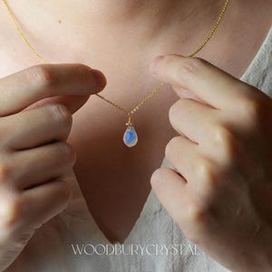 May include: A delicate gold chain necklace with a teardrop-shaped opal pendant. The opal is a milky white color with a hint of blue.