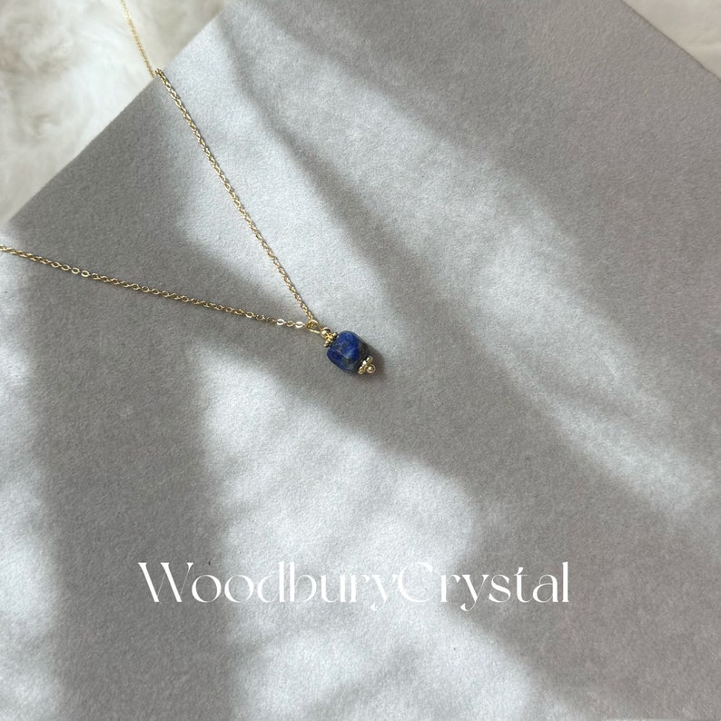 Peut inclure: Un collier en cha&icirc;ne dor&eacute;e avec un pendentif en lapis-lazuli bleu fonc&eacute;. La pierre est de forme irr&eacute;guli&egrave;re naturelle. Le collier est sur un fond blanc avec le texte "Woodbury Crystal" en bas.