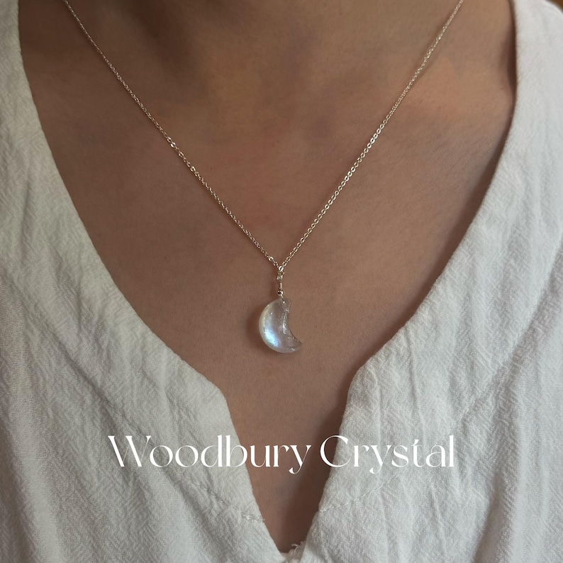 May include: A silver chain necklace with a crescent moon pendant made of iridescent moonstone. The pendant is a light blue color with a white sheen. The necklace is on a white background with the text "Woodbury Crystal" in white.