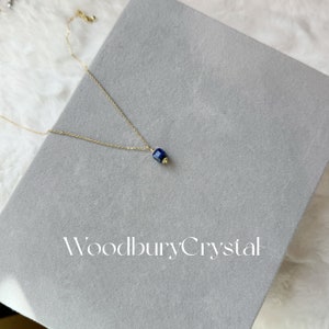 Peut inclure: Un collier en cha&icirc;ne dor&eacute;e avec un pendentif en pierre pr&eacute;cieuse bleu fonc&eacute;. Le pendentif est de forme carr&eacute;e avec des bords arrondis. Le collier est sur une surface grise avec le texte "Woodbury Crystal" imprim&eacute; dessus.