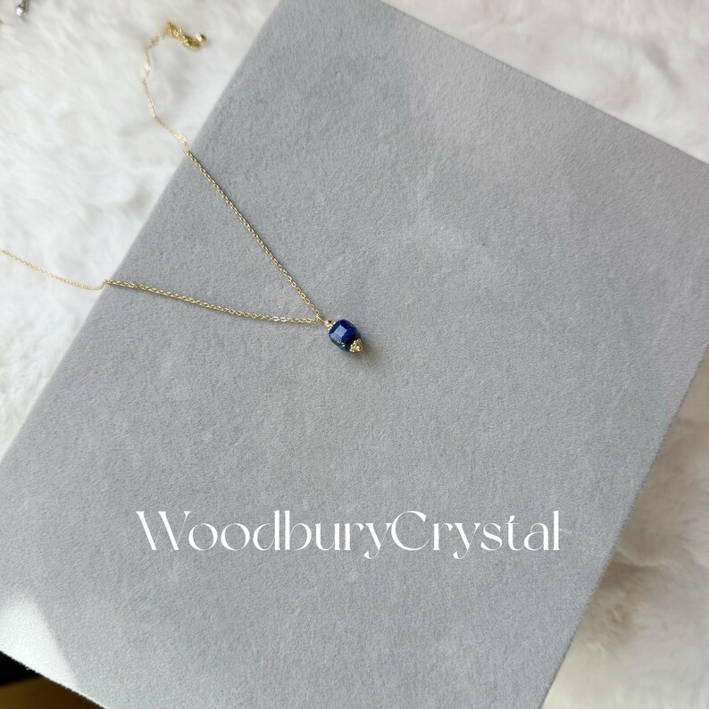 Peut inclure: Un collier en cha&icirc;ne dor&eacute;e avec un pendentif en pierre pr&eacute;cieuse bleu fonc&eacute;. Le pendentif est de forme carr&eacute;e avec des bords arrondis. Le collier est sur une surface grise avec le texte "Woodbury Crystal" imprim&eacute; dessus.